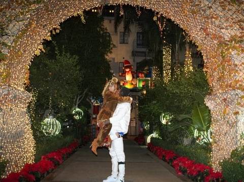 Woman in white outfit posing under illuminated archway decorated with greenery, red flowers, and hanging ornaments