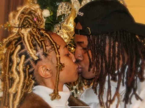 Two people with blonde and dark dreadlocks kissing, wearing white shirts and festive gold decorations