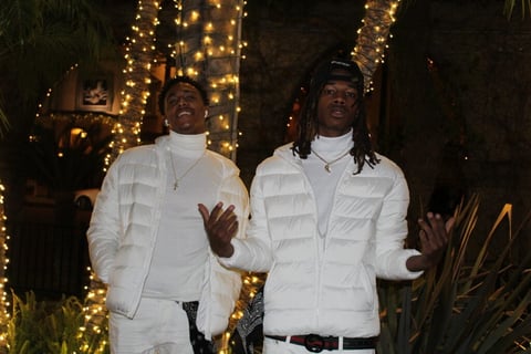 Two men in white puffer jackets pose together at night surrounded by illuminated trees and warm golden lights