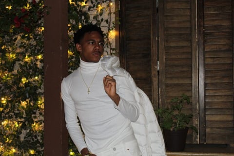 Man in white turtleneck and white jacket posed against holiday decorated backdrop with illuminated garland