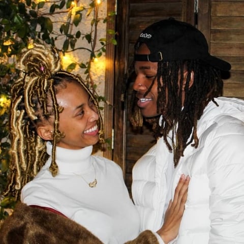 Two people with dreadlocks wearing white outfits smiling at each other indoors near decorative gold and green foliage